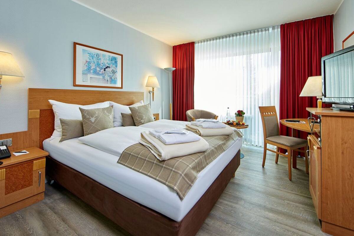 A hotel room with a large bed, a television, a desk with a chair, and a window with red curtains.
