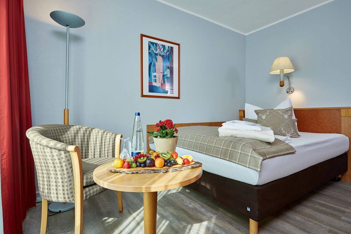 A hotel room with a bed, a chair, a round wooden table with fruit, a lamp, and a painting on the wall.