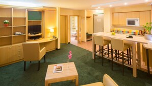 Une chambre d’hôtel compacte comprenant un coin cuisine, un espace salon avec un canapé et une petite table, une télévision et une salle de bain visible à travers une porte ouverte.