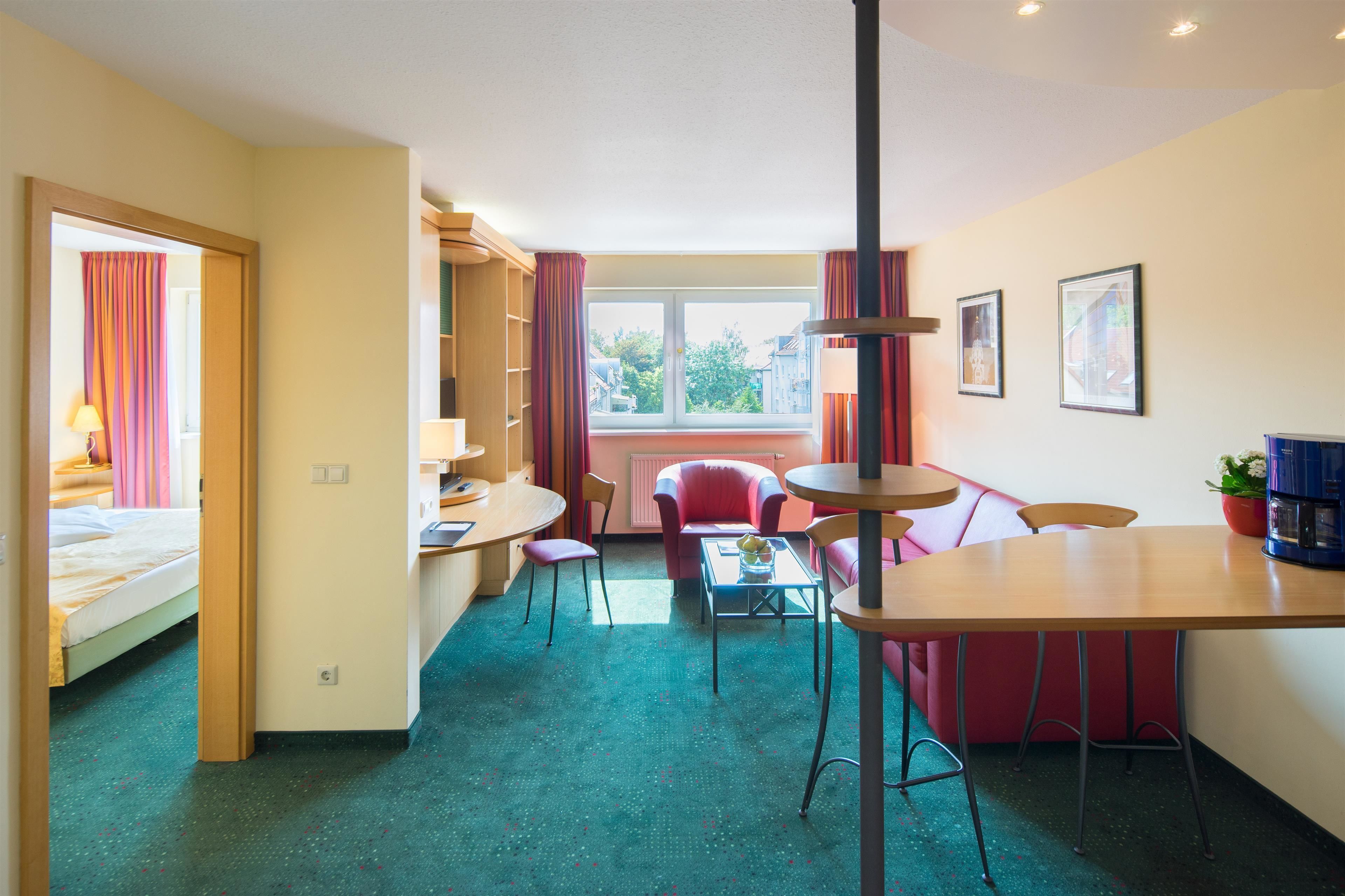 A hotel room with a bed, a desk, a dining table, chairs, a sofa, a window, and a radiator.