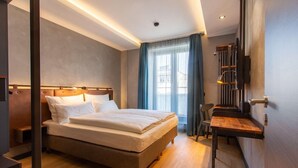 A modern hotel room with a large bed, a desk, and a view of the city through large windows. - Golden Leaf Parkhotel Im Lehel (Munich)