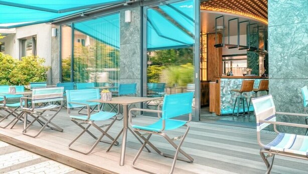 An outdoor seating area with blue chairs and tables, a glass door entrance, and a kitchen visible inside. - Golden Leaf Parkhotel Im Lehel (Munich)