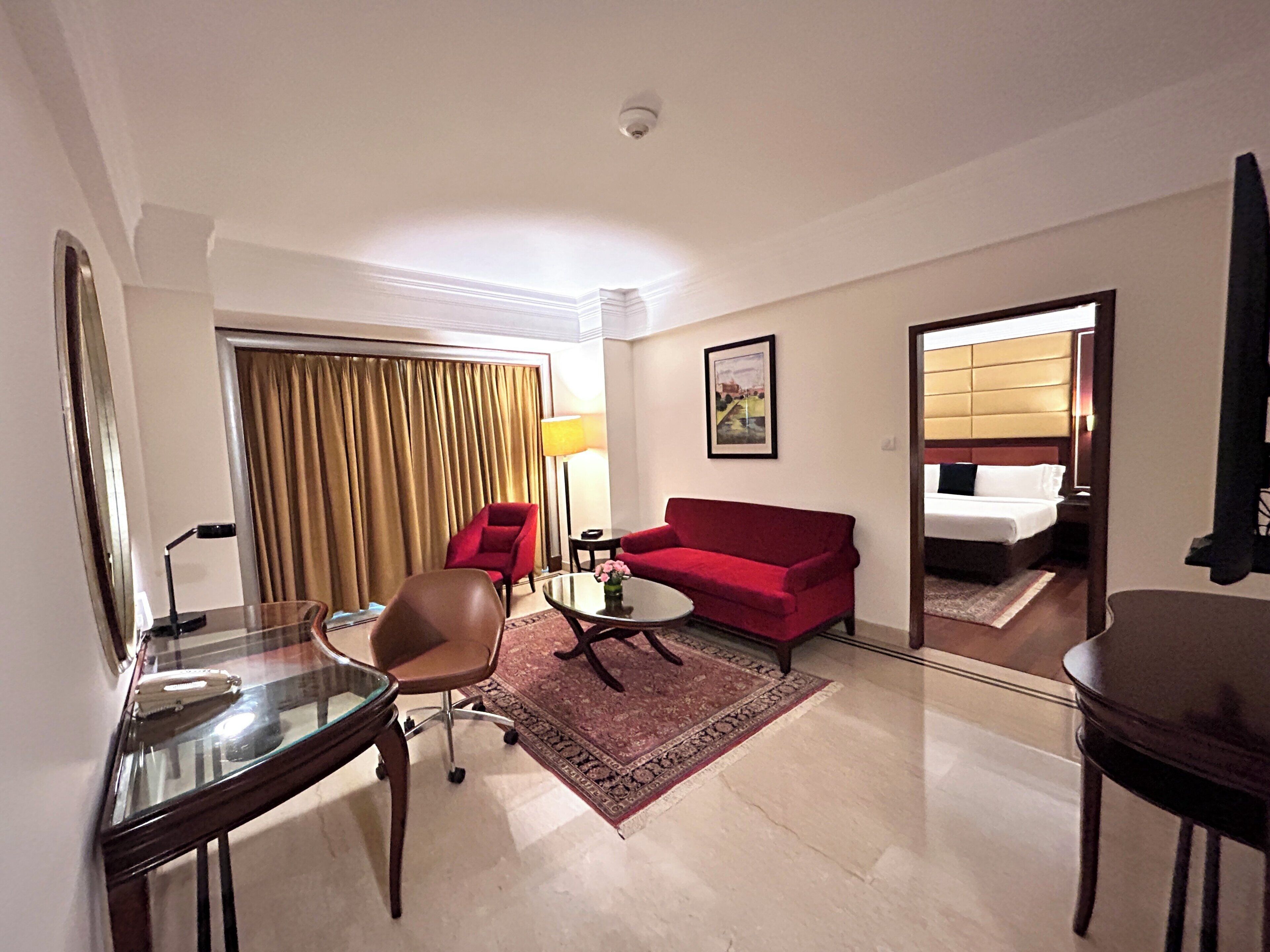 Une chambre d’hôtel avec un lit, un canapé rouge, une table basse en verre, un fauteuil marron et un bureau avec une lampe.
