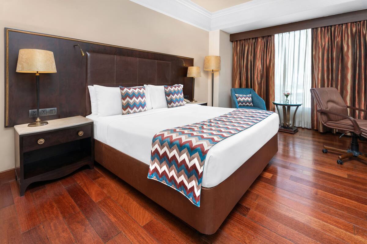A hotel room with a large bed, two bedside tables, a chair, a small table, and a window with curtains.