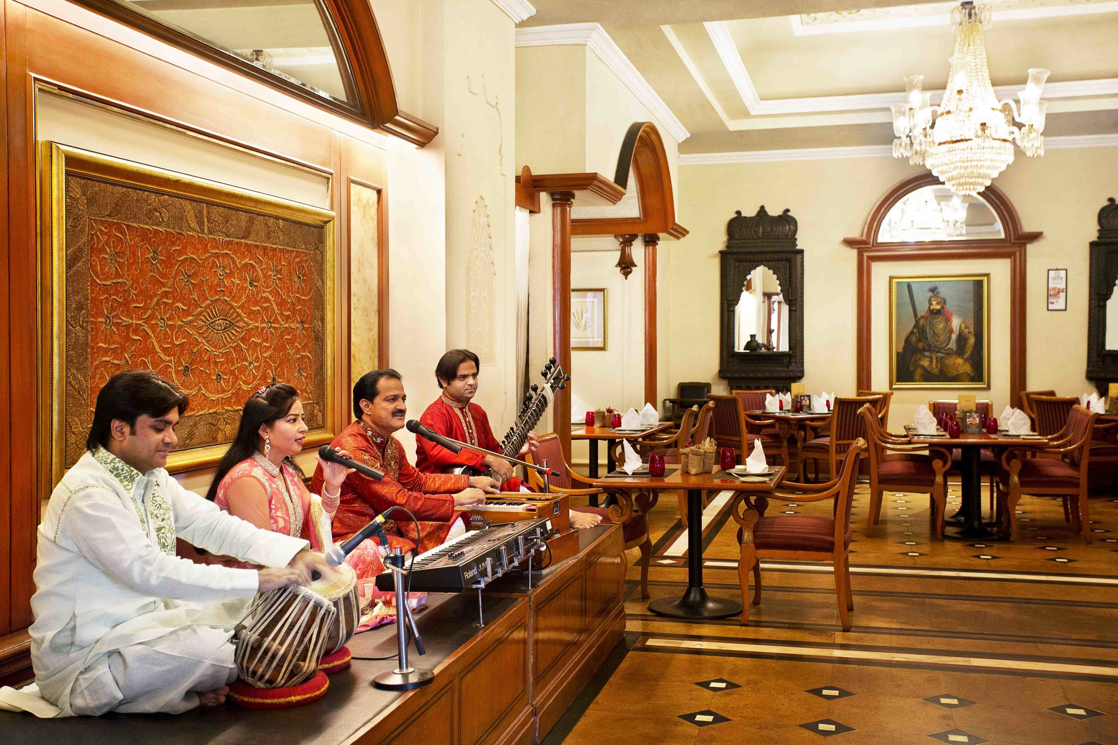 A room with a group of musicians performing traditional instruments. There are wooden tables and chairs, a chandelier, and framed artwork on the walls.