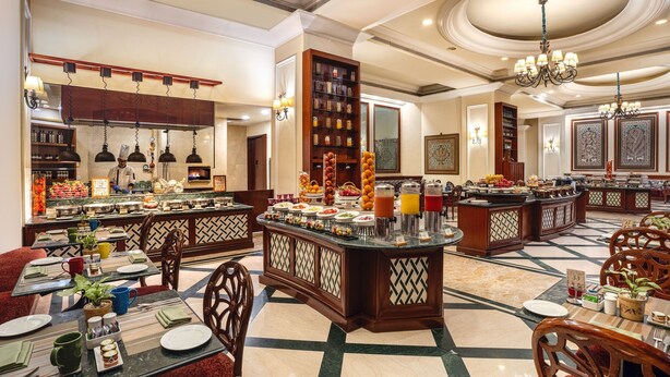 A hotel dining area with multiple seating arrangements, a buffet setup, and a kitchen in the background.