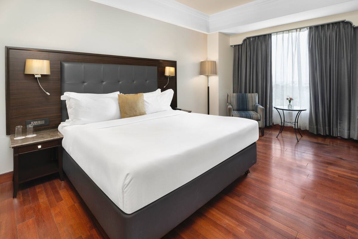 A hotel room with a large bed, two bedside lamps, a small table with a vase, a chair, and a view of the outdoors through curtains.