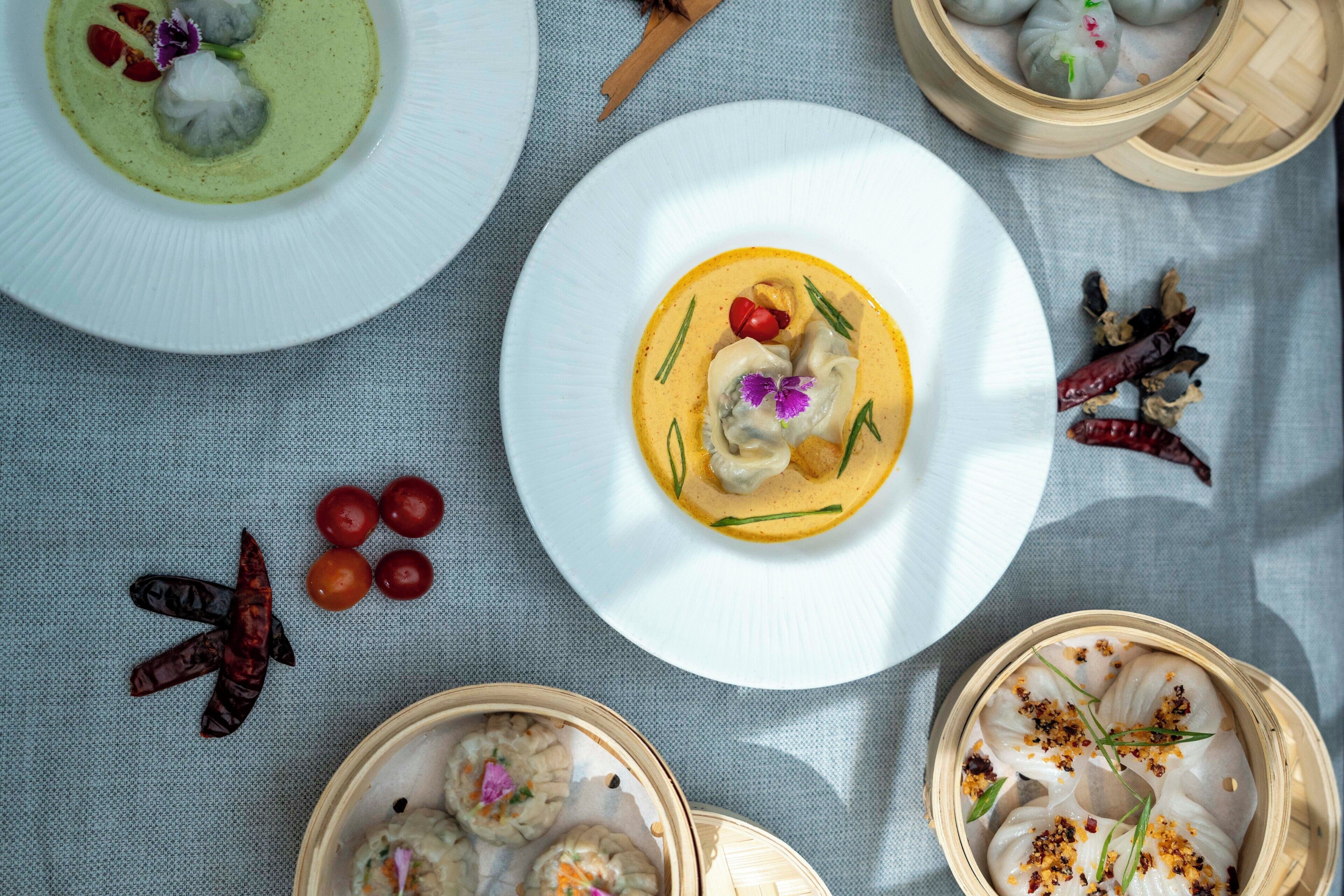 A table with various dishes, including dumplings in bamboo steamers, a plate with a green sauce, and a plate with a yellow sauce and vegetables.
