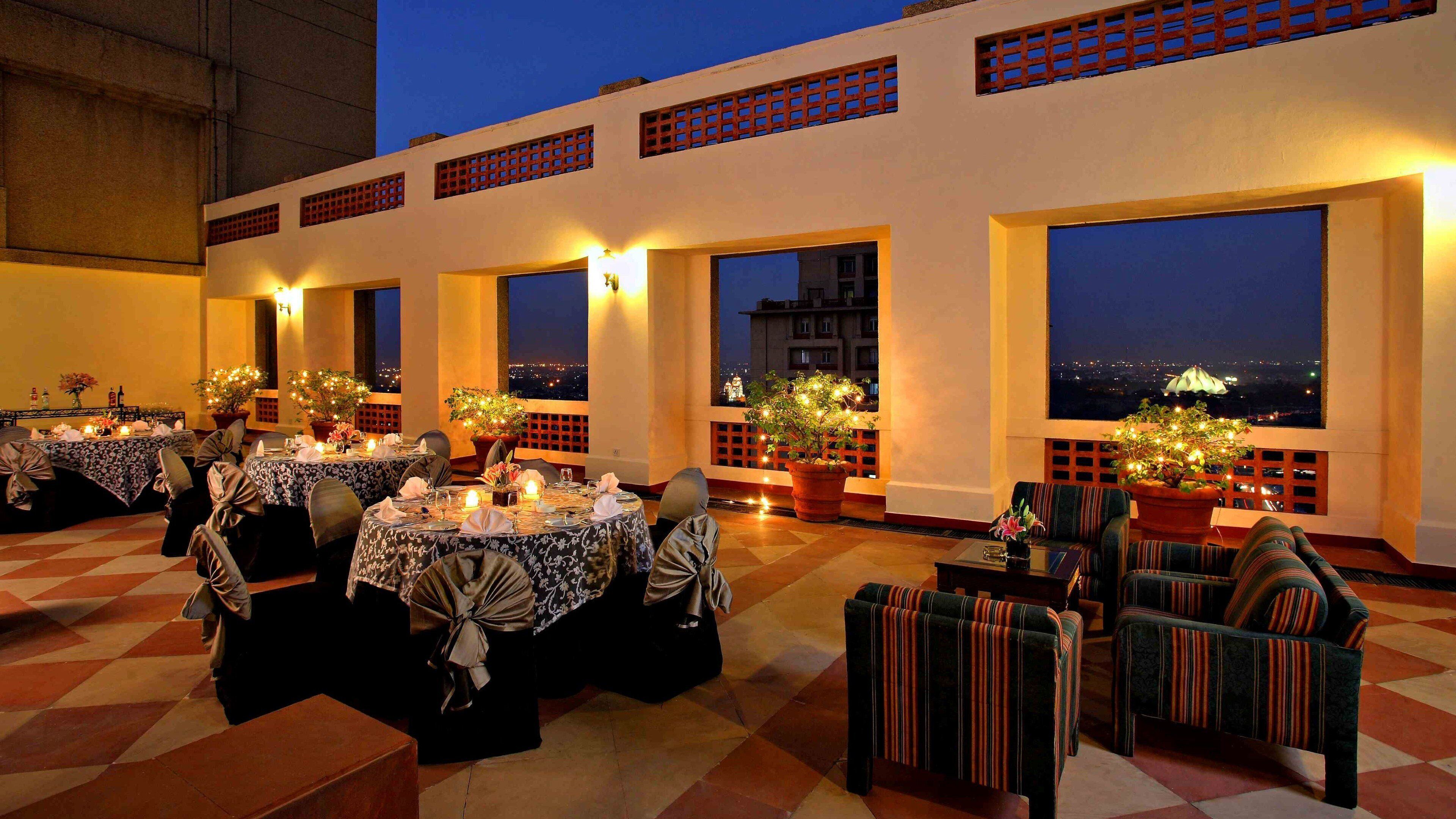 A rooftop dining area with tables set for a meal, potted plants, and a view of the cityscape at night.