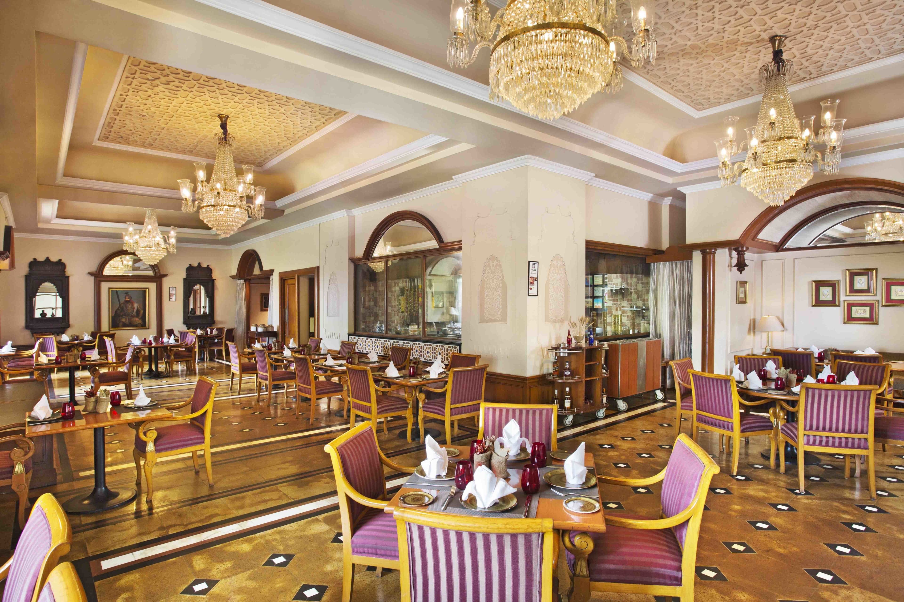 A spacious hotel dining area with multiple tables and chairs, ornate chandeliers, and classic decor.