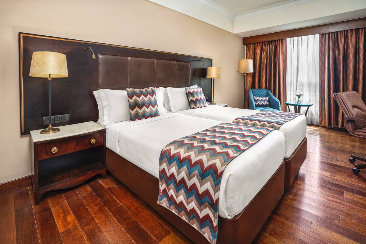 A hotel room with a large bed, two bedside tables, a chair, a small table, and a window with curtains.