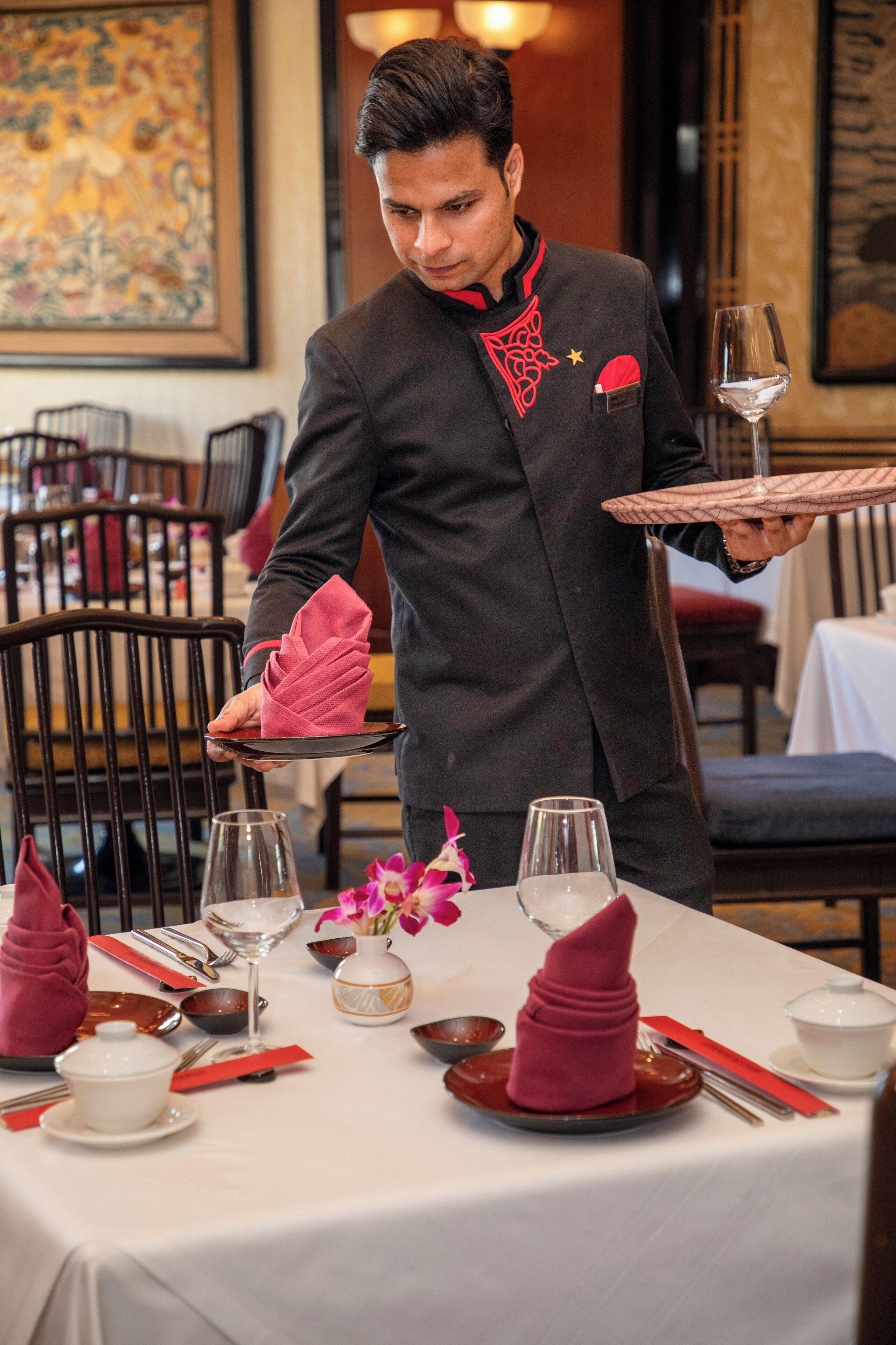 A waiter serving a table with a white tablecloth, red napkins, and a vase of flowers.