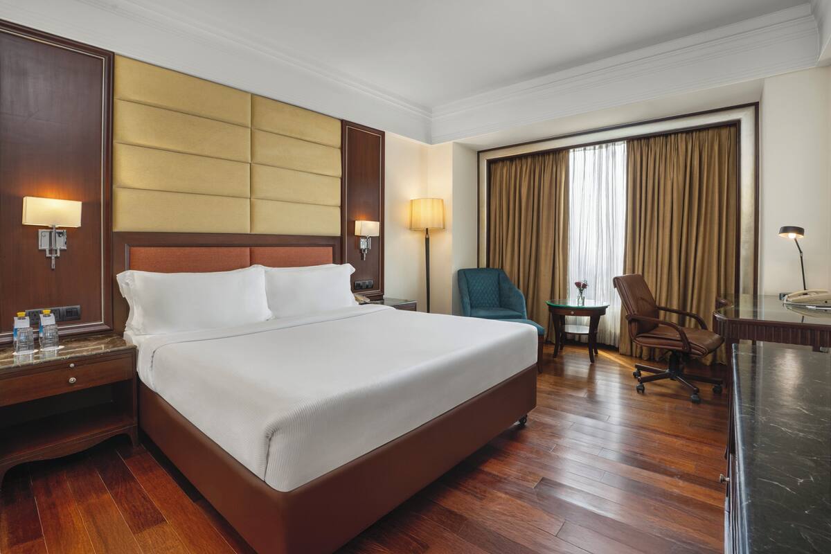 A hotel room with a large bed, a desk, a chair, a lamp, and a window with curtains.