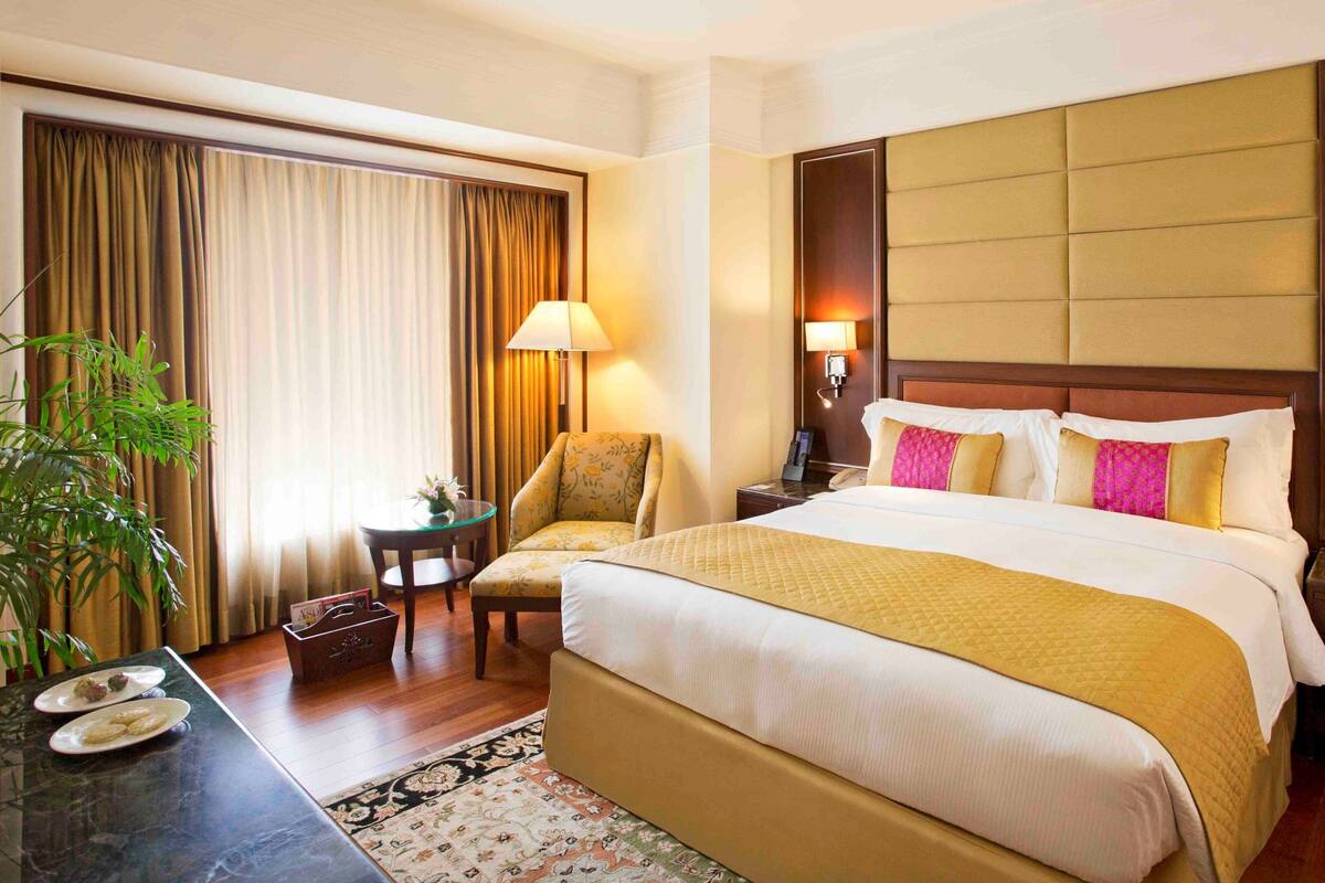 A hotel room with a large bed, a bedside table, a chair, a desk with a lamp, and a window with curtains.
