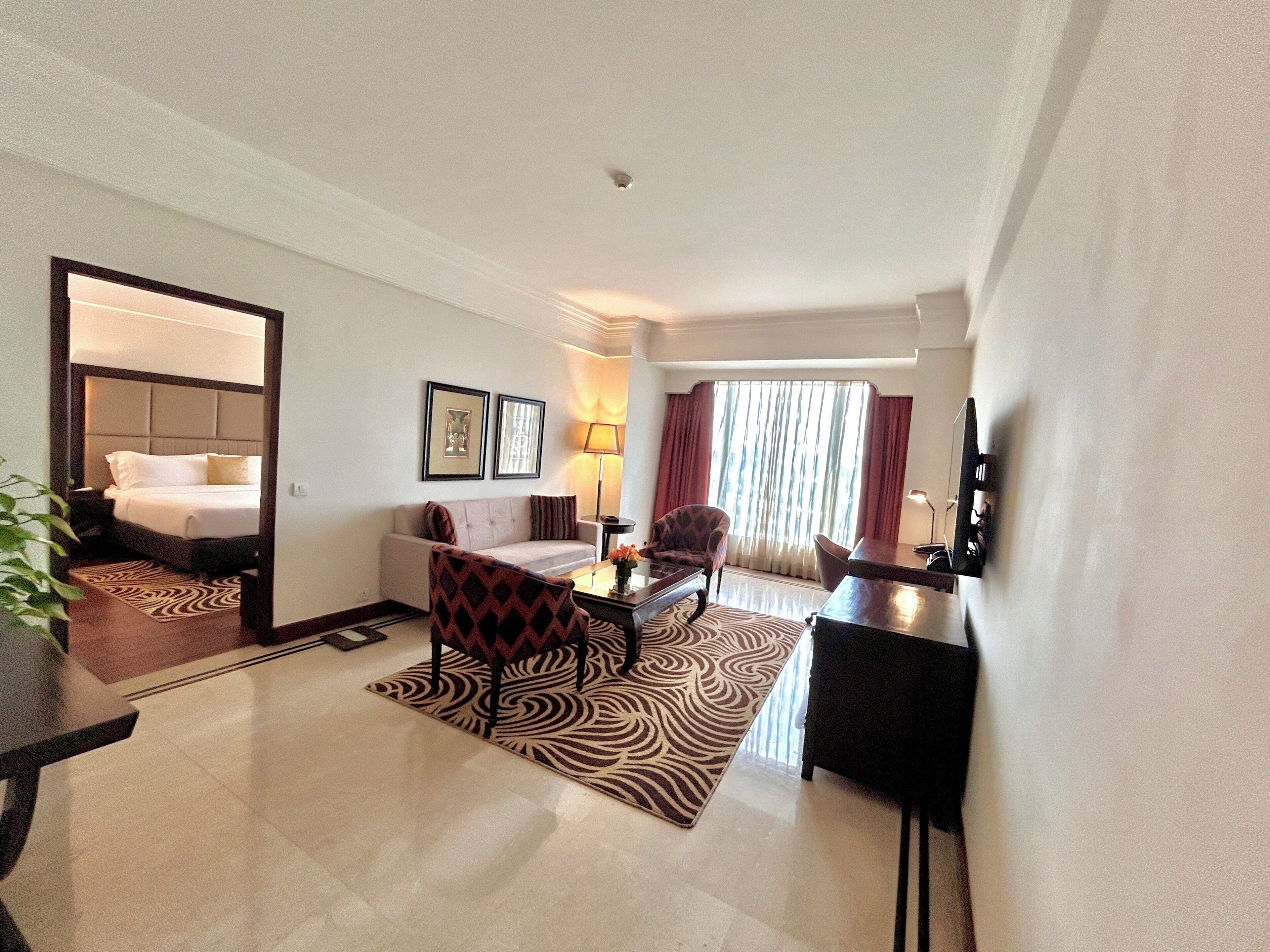 A hotel room with a bed, a sofa, a dining table, and a chair. There is a mirror on the wall and a window with curtains.