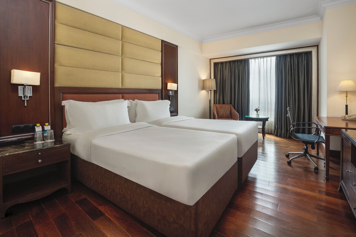 Une chambre d’hôtel avec deux lits, un bureau, une chaise, une lampe et une fenêtre avec des rideaux.