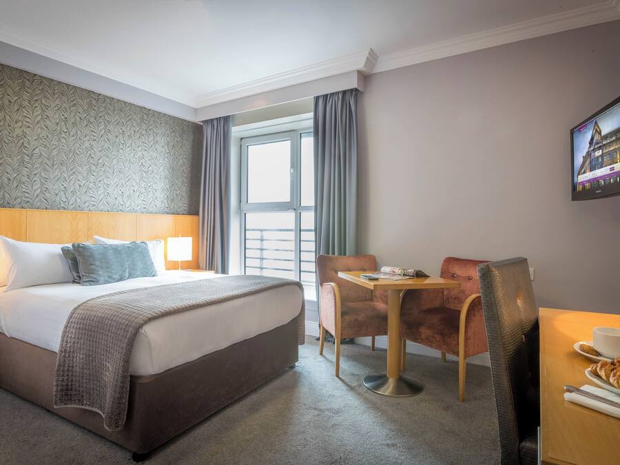 Una habitación de hotel con cama, escritorio, silla, televisión y ventana con cortinas.