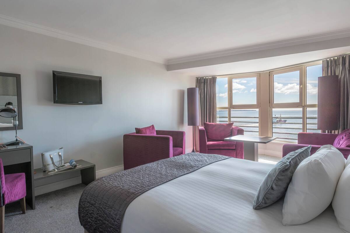A hotel room with a bed, a desk, a chair, a TV, and a large window with a view of the sea.