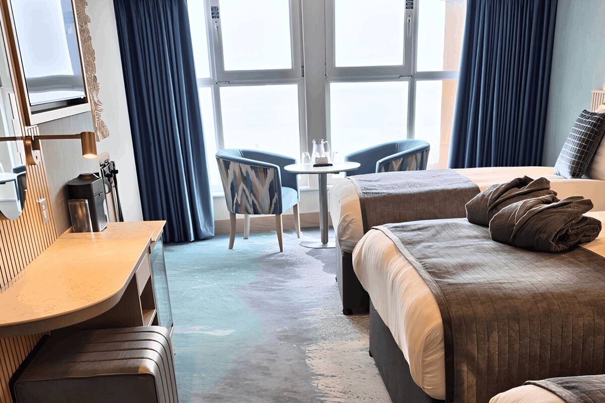A hotel room with two beds, a desk, a chair, a small table, and a large window.
