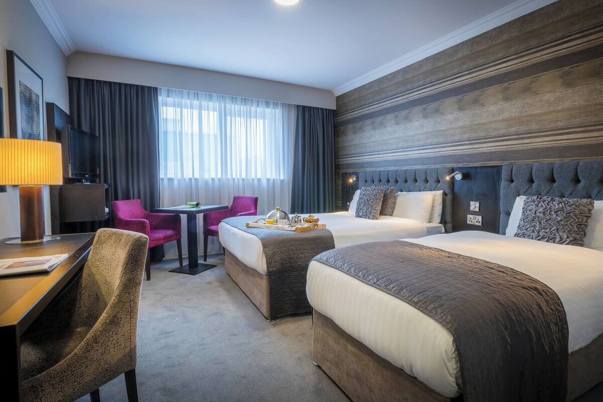 A hotel room with two beds, a desk with a lamp, a chair, a table with a tray of breakfast, and a large window with curtains.