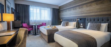 Quarto de hotel com duas camas, uma mesa com luminária, uma cadeira, uma mesa com bandeja de café da manhã e uma janela grande com cortinas.