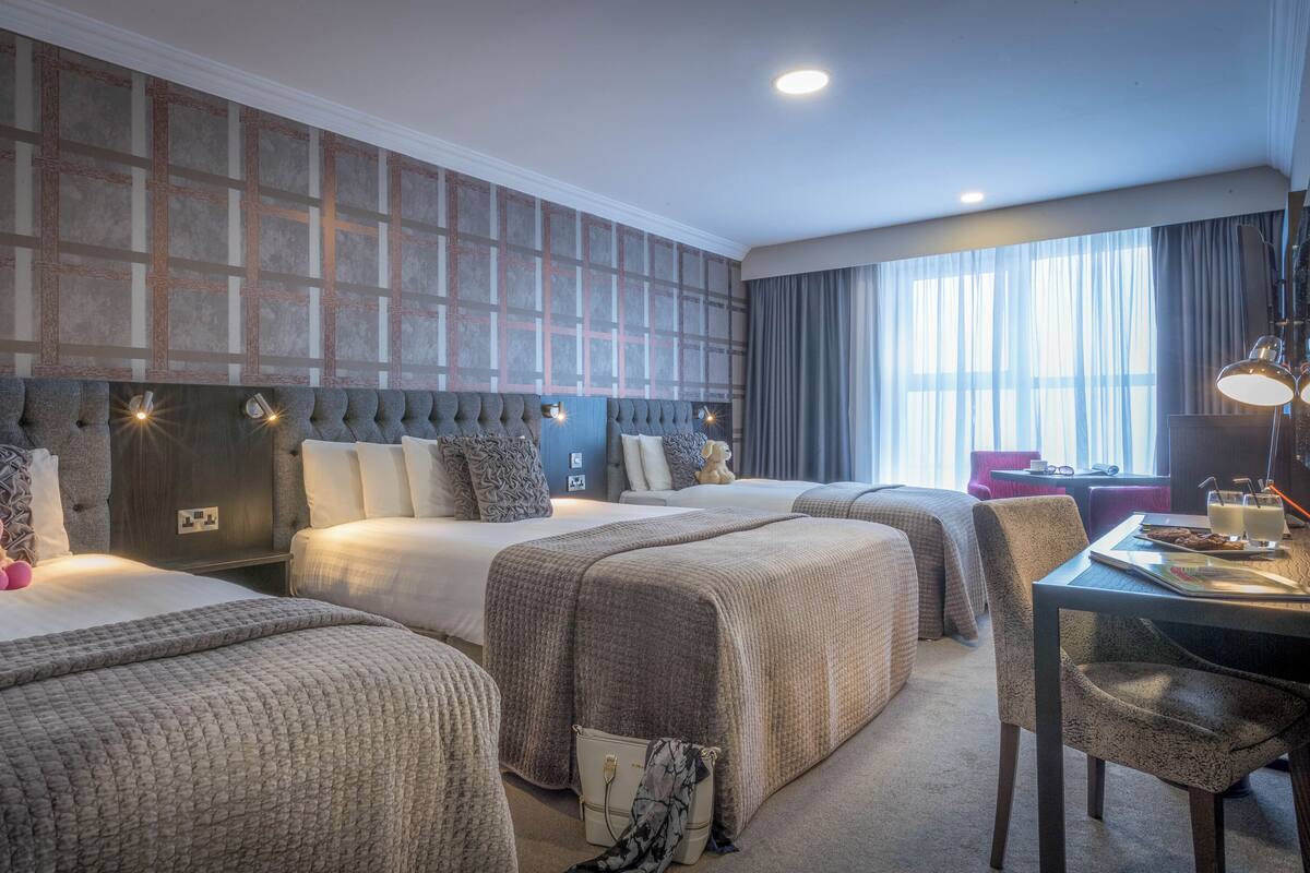 A hotel room with three beds, a desk with a chair, a lamp, and a view of the outdoors.