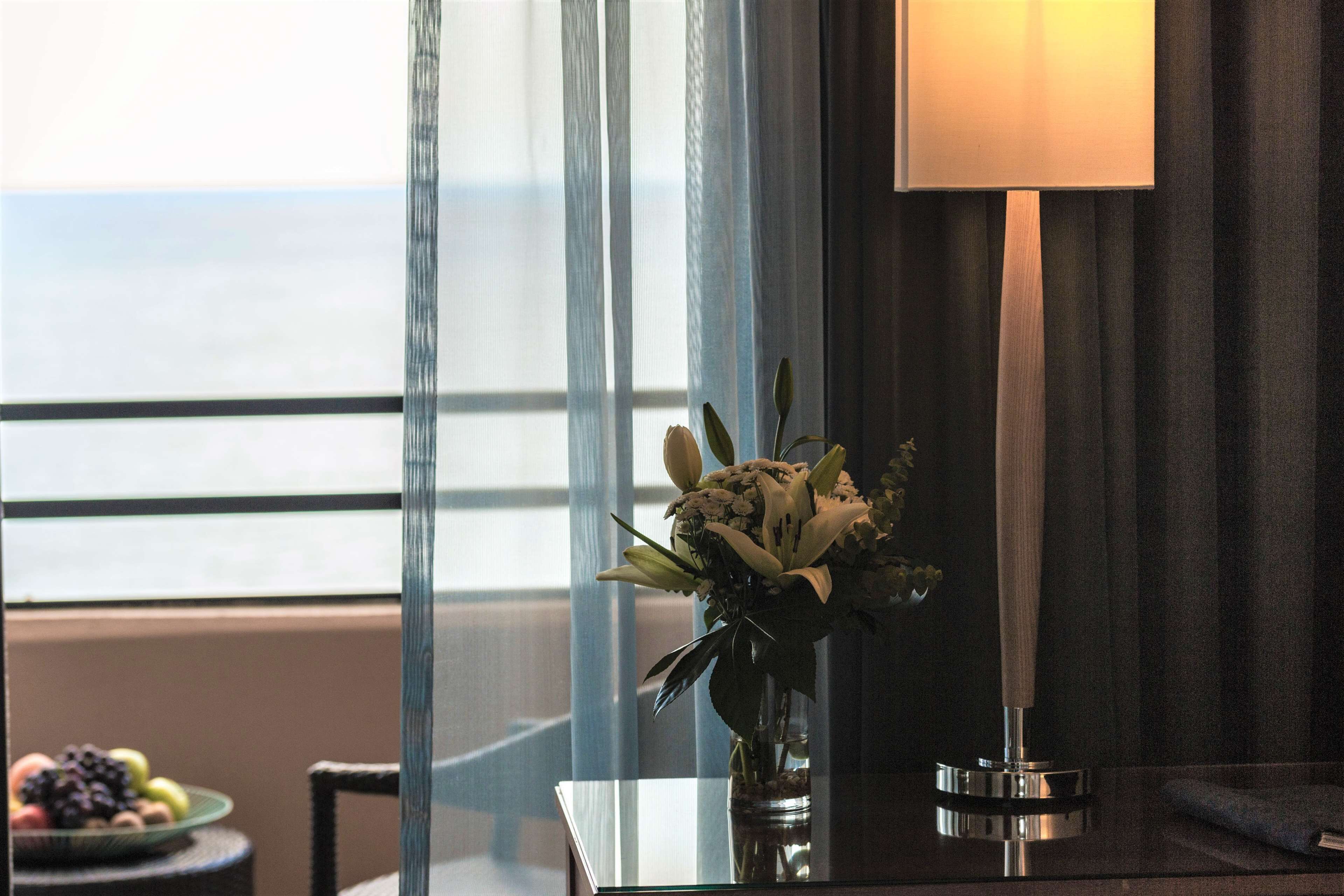 A room with a view of the sea, a vase with flowers on a table, and a lamp.