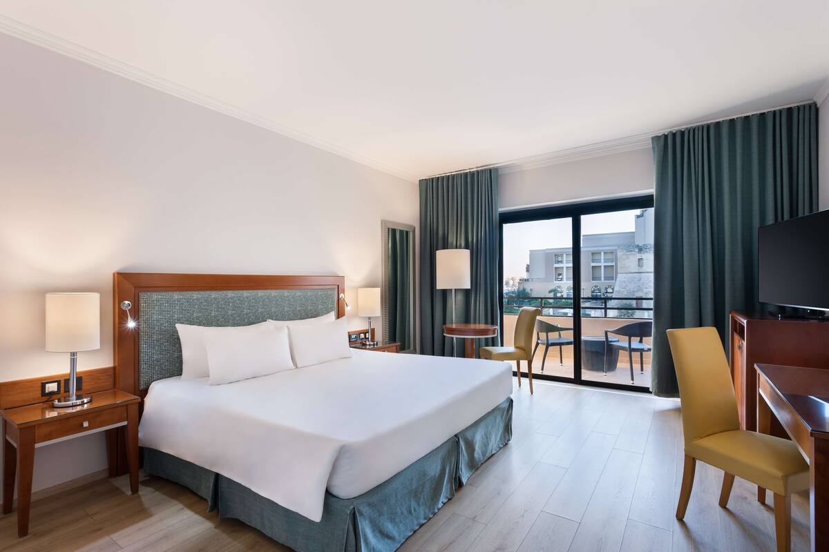 A hotel room with a large bed, two bedside tables, a TV, a desk, and a balcony with a view of buildings.