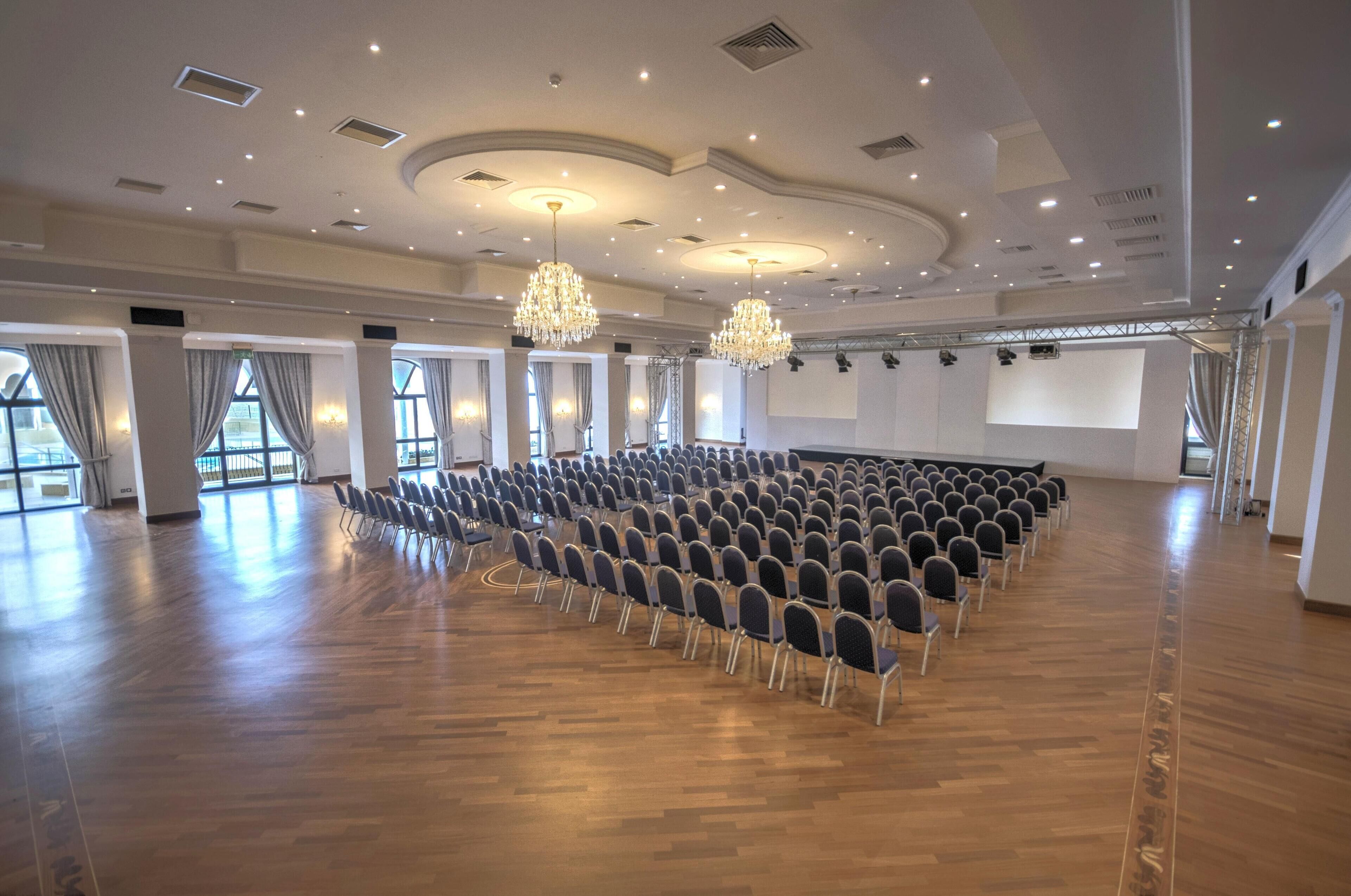 Een grote congreszaal met rijen stoelen, een podium en een groot scherm.