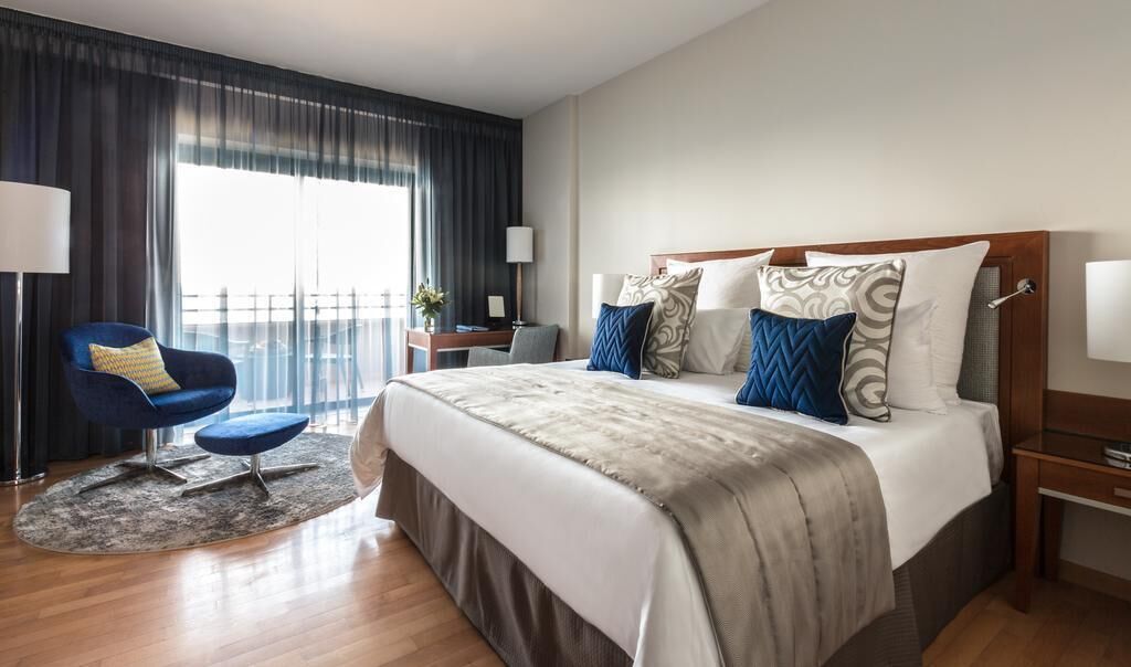 A hotel room with a large bed, a blue armchair, a desk, and a view of the outdoors.