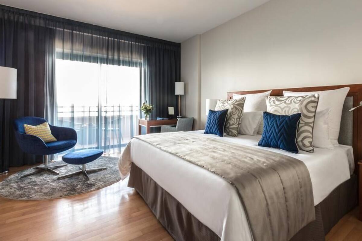 A hotel room with a large bed, a blue armchair, a desk, and a view of the outdoors.