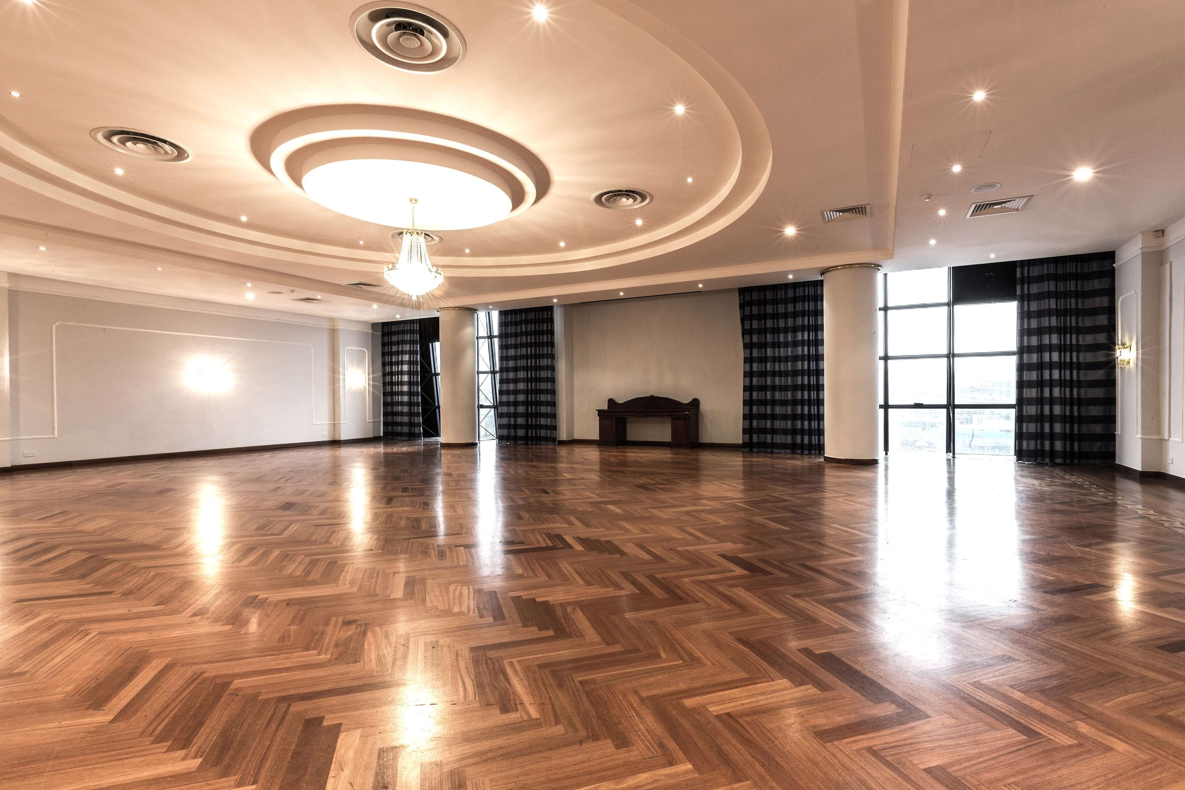 A spacious room with a wooden parquet floor, a central chandelier, and large windows with curtains.