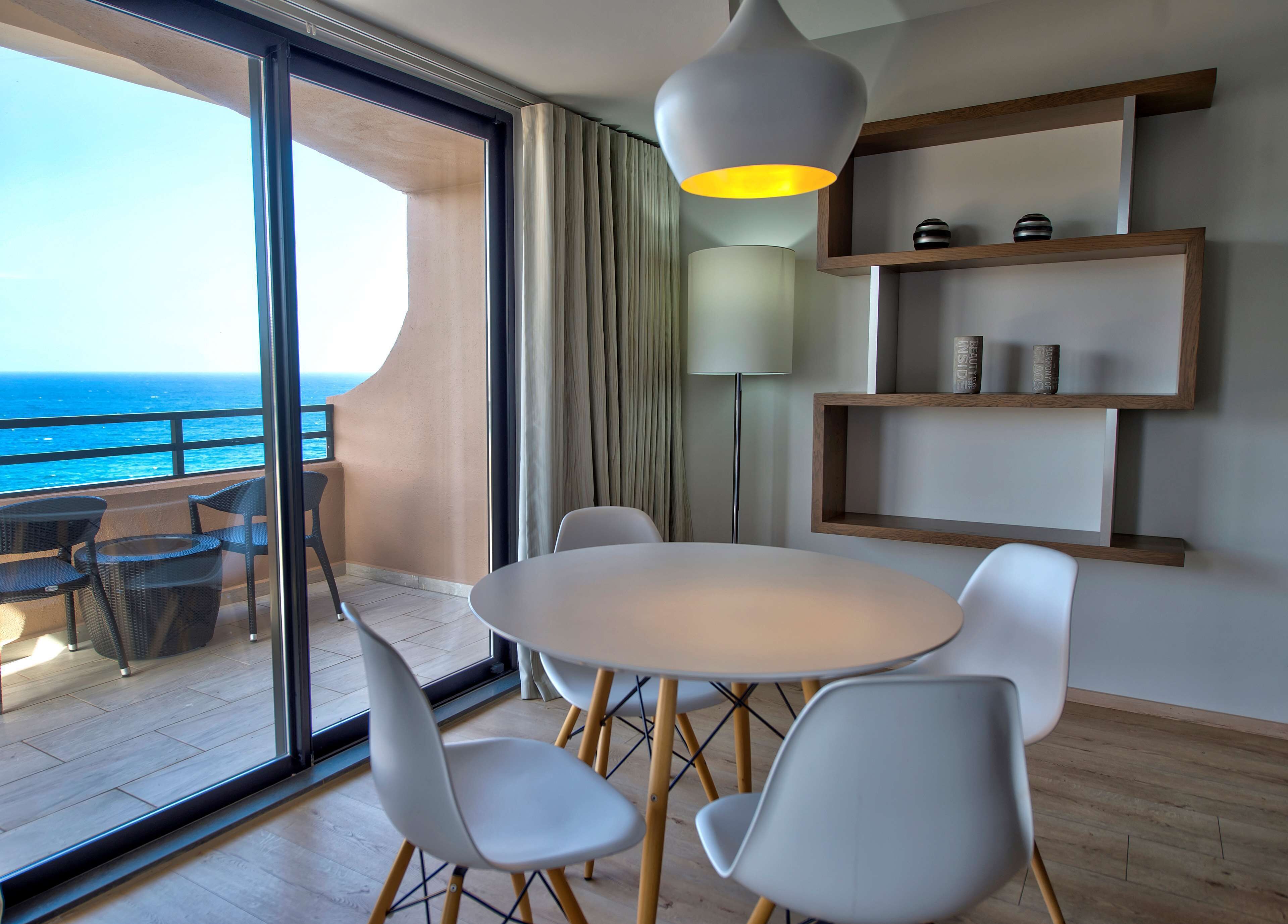 Una habitación moderna con balcón con vistas al océano, que incluye una mesa redonda con sillas, una lámpara de pie y estanterías de madera con objetos decorativos.