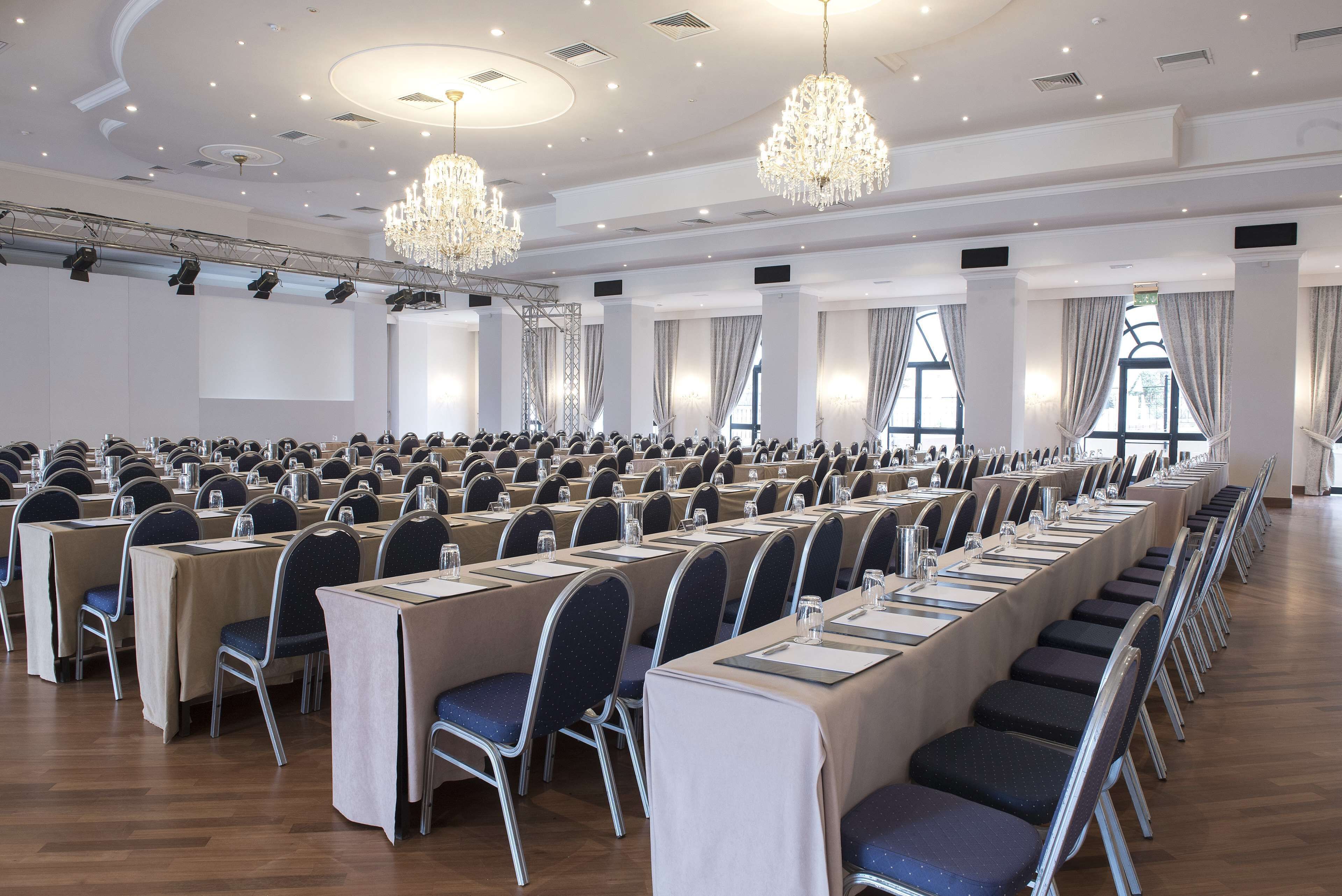 A conference room with rows of tables and chairs, large windows, and chandeliers.