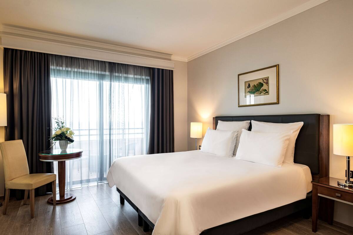 A hotel room with a large bed, a small round table with a vase of flowers, a chair, a nightstand with a lamp, and a framed picture on the wall.