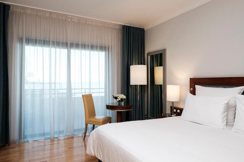 A hotel room with a bed, a chair, a table with a vase, and a large window with curtains.