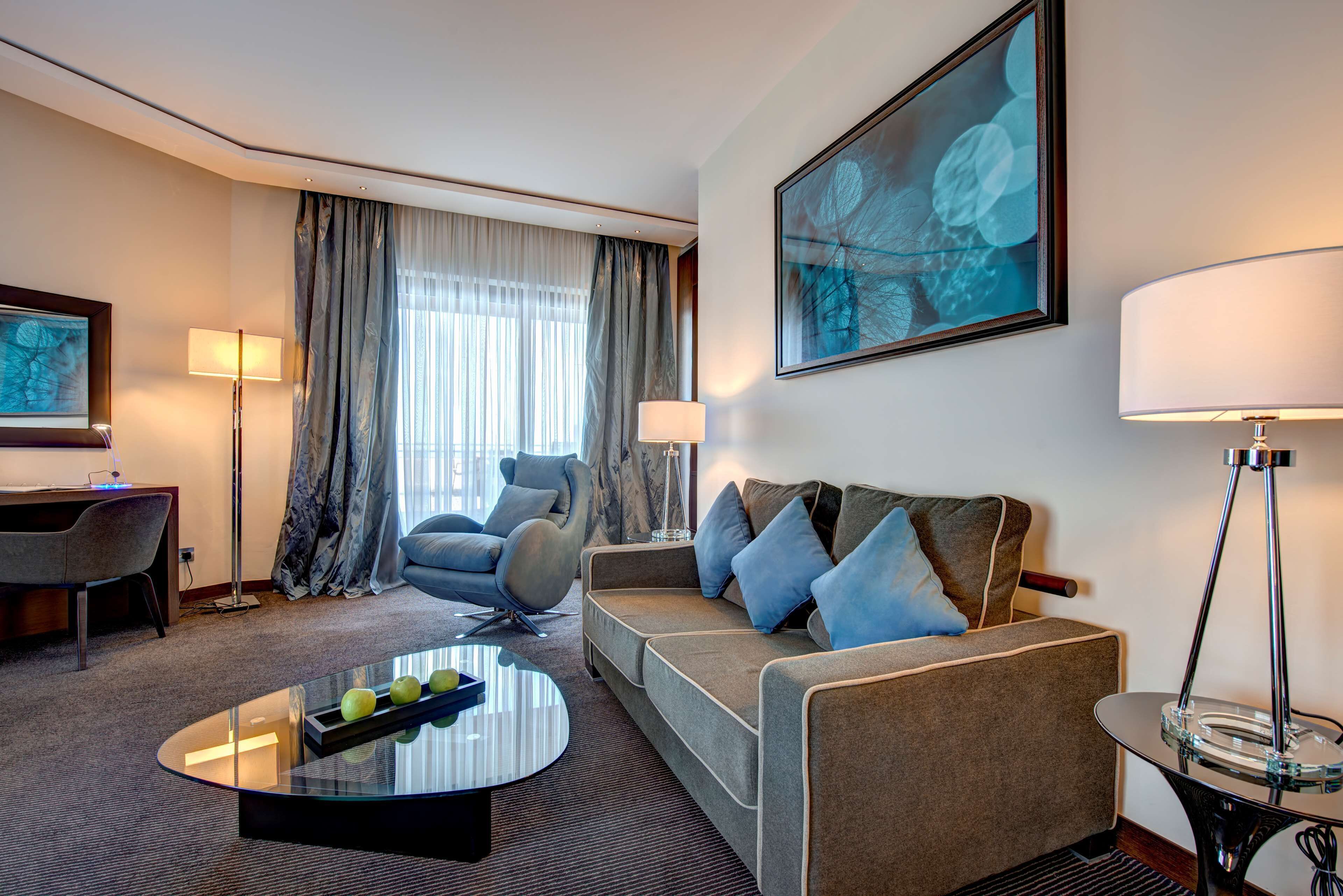 A modern hotel room with a sofa, a chair, a desk, and a coffee table with apples.