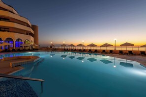A swimming pool with lounge chairs and umbrellas, surrounded by a building with balconies and a sunset view.