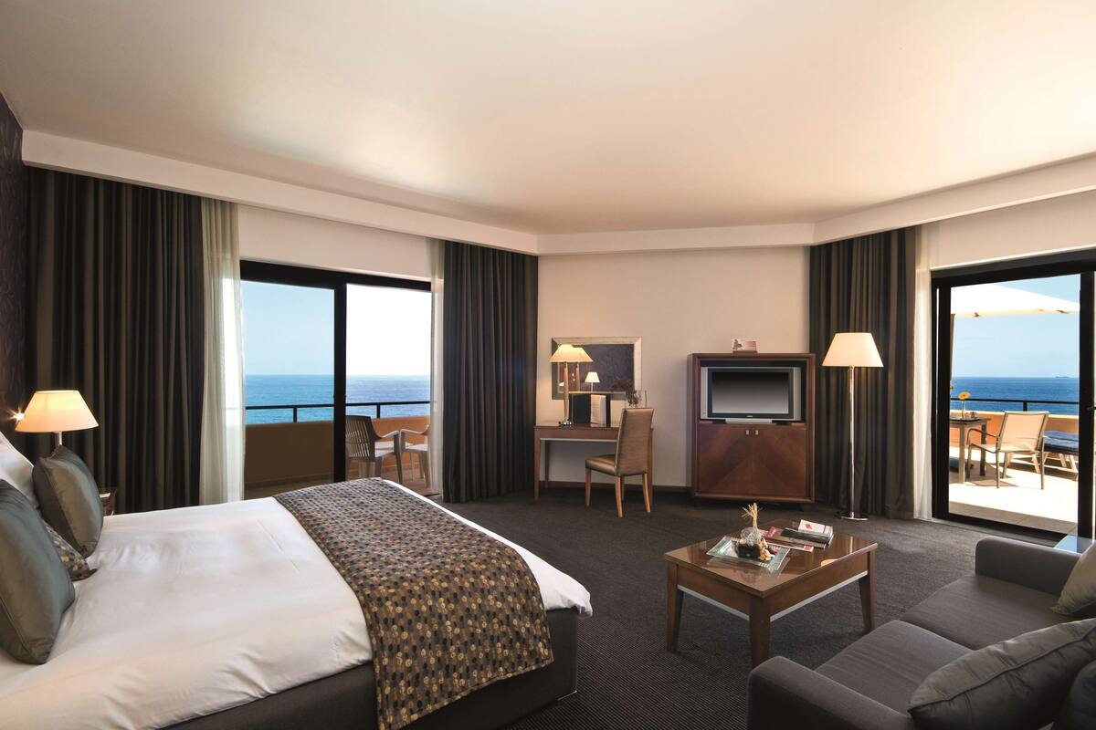 A hotel room with a large bed, a desk with a TV, a sofa, and a balcony with a view of the ocean.