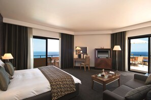 Habitación de hotel con una cama grande, un escritorio con televisor, un sofá y un balcón con vista al océano.