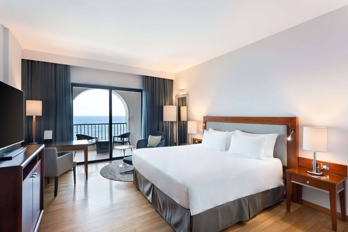 A hotel room with a large bed, a desk, a chair, and a view of the sea.