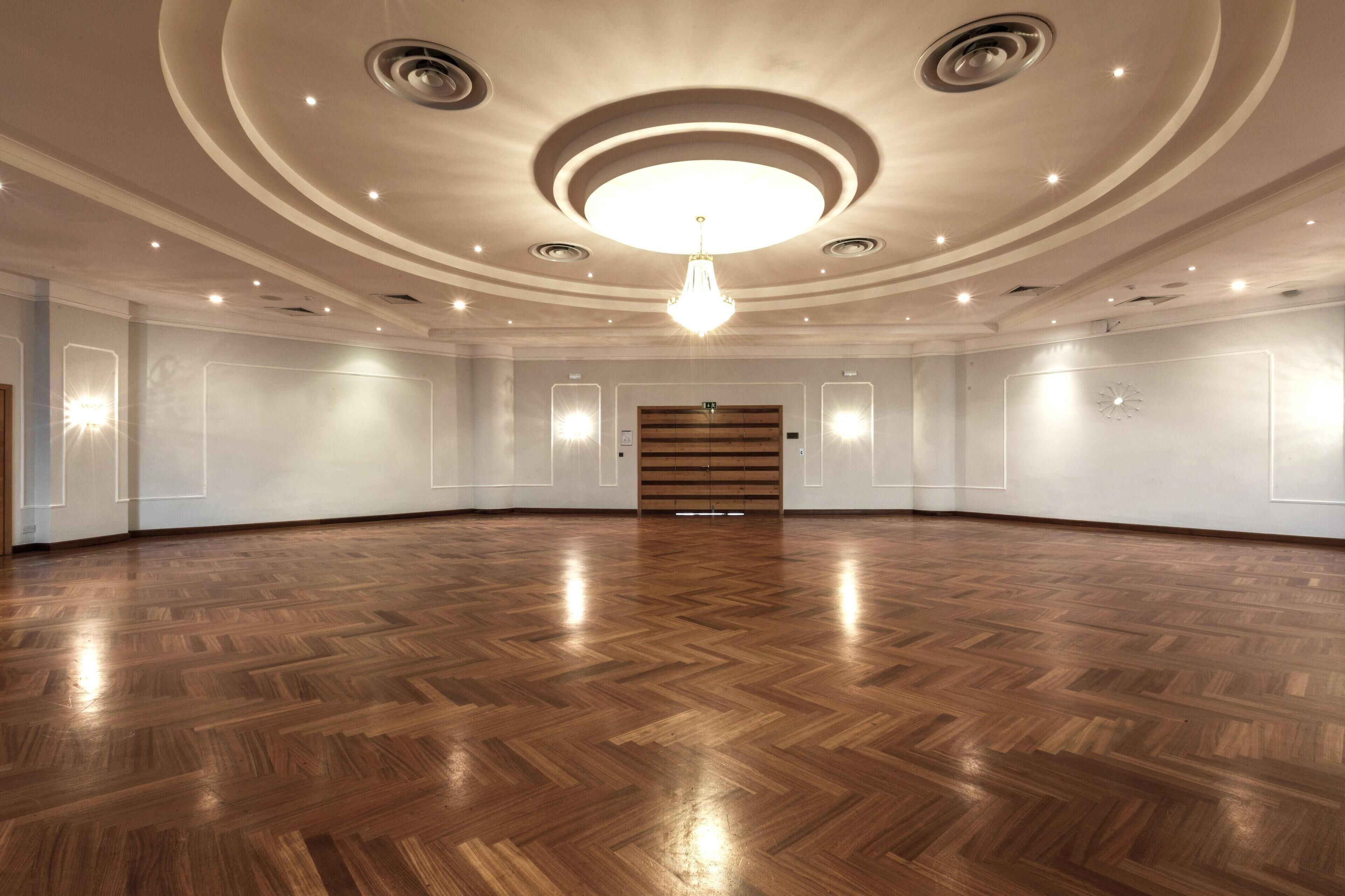 A spacious, well-lit room with a wooden parquet floor, a central chandelier, and a wooden door at the far end.