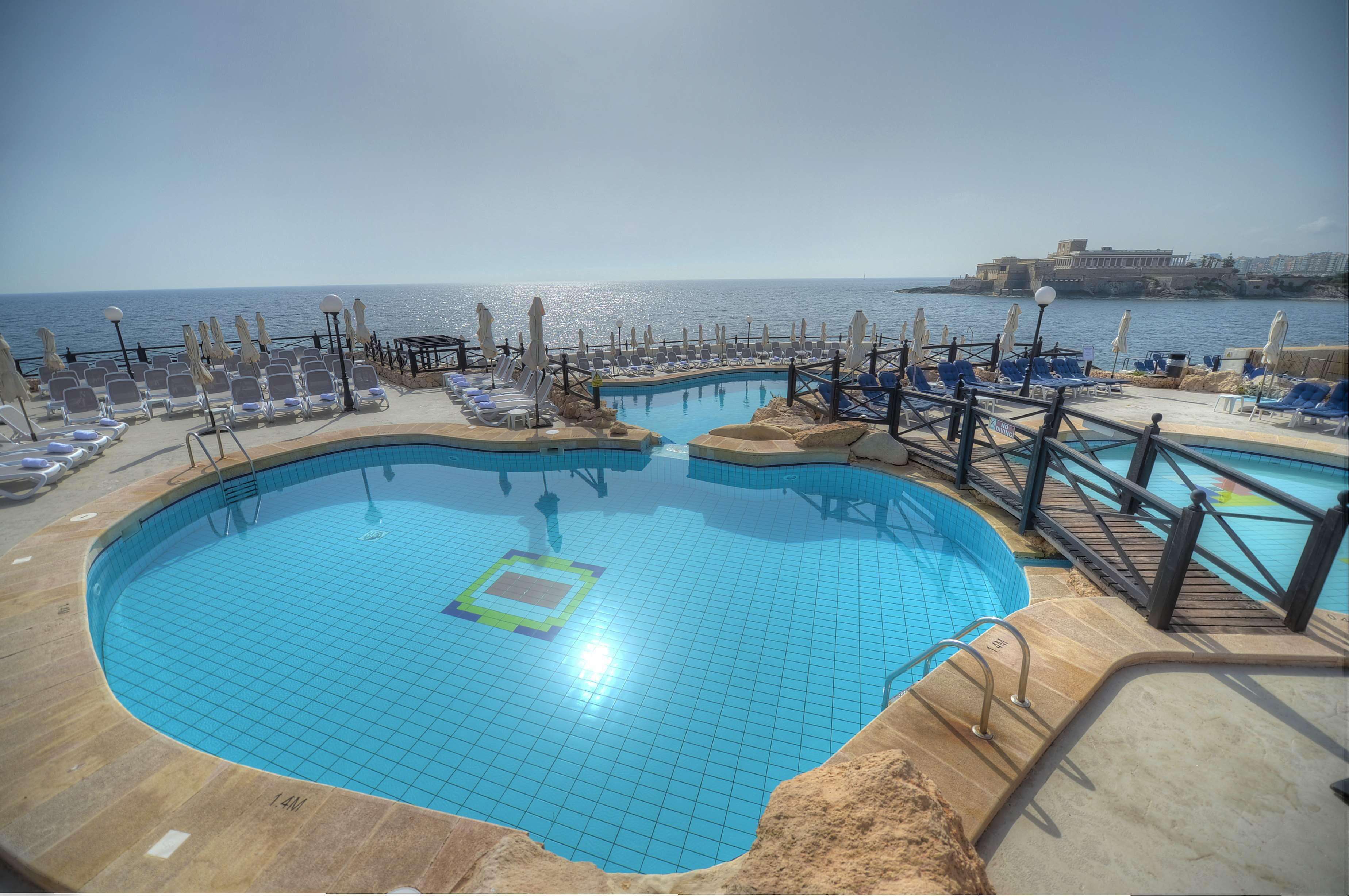 A pool area with sun loungers and umbrellas, a clear view of the sea, and a historic building in the background.
