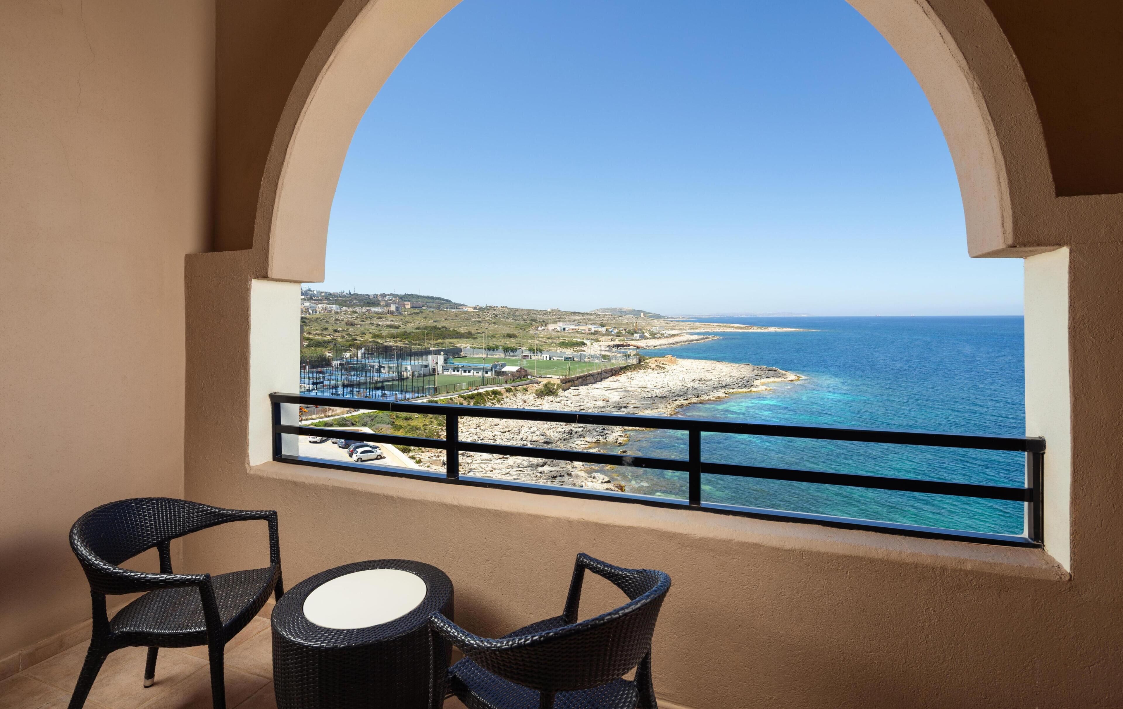 A balcony with wicker chairs and a table, overlooking a coastal view with clear blue sea and a rocky shoreline.