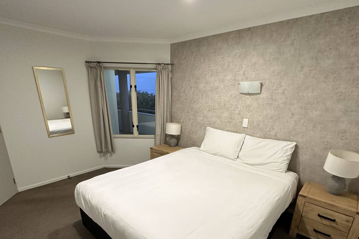 Un grand lit, bien fait, dans une chambre d’hôtel, avec une fenêtre, un miroir et deux tables de chevet équipées de lampes.