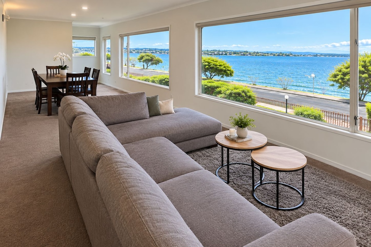 Un vaste espace de vie comprenant un grand canapé d’angle, une table à manger avec des chaises et une vue sur la mer grâce à de grandes fenêtres.