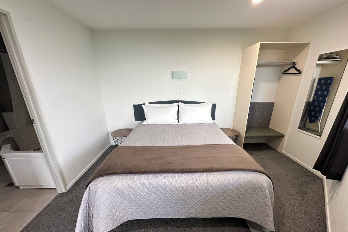 Une chambre d’hôtel bien rangée, avec un grand lit, une petite table de chevet, une chaise, une armoire et un miroir.