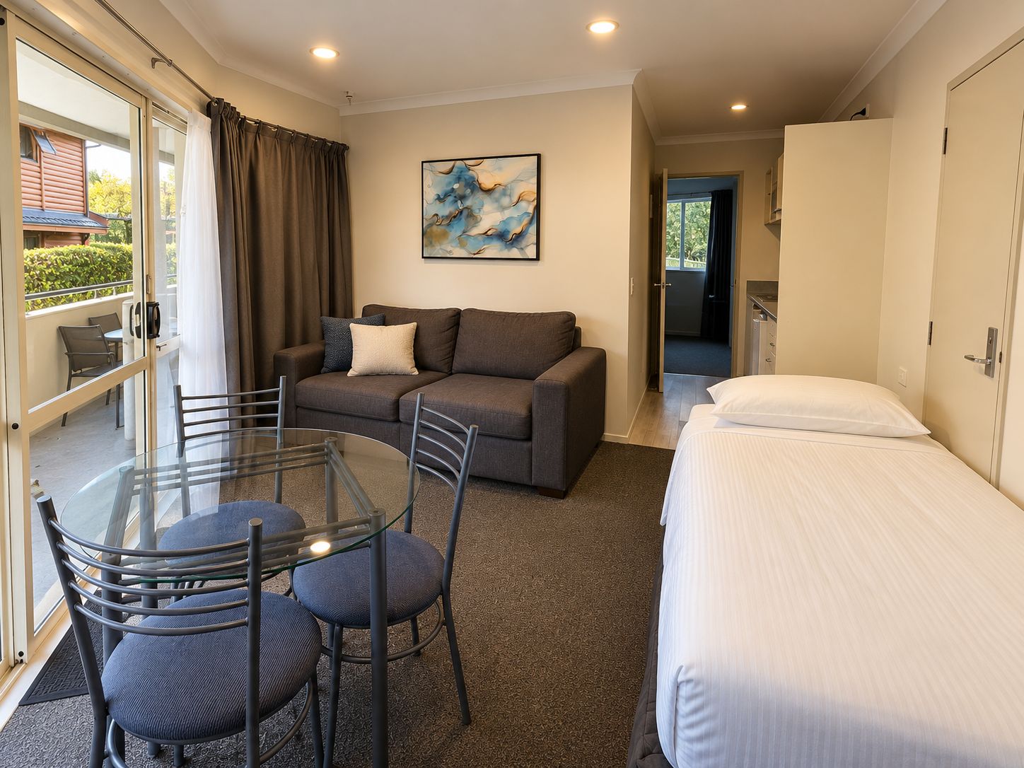A hotel room with a glass table, chairs, a sofa, a bed, and a painting on the wall.