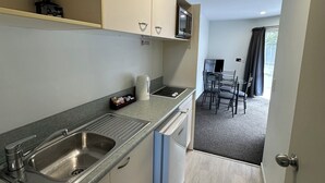 Een compacte keuken met een ingebouwde magnetron, een spoelbak en een waterkoker. Een eethoek met een tafel en stoelen is zichtbaar door een geopende deur.