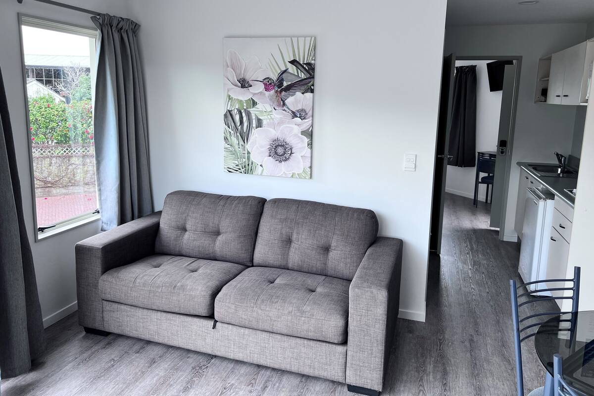 Un salon moderne avec un canapé gris, un coin repas avec une table ronde et des chaises, une cuisine avec des meubles blancs et une grande fenêtre avec des rideaux.